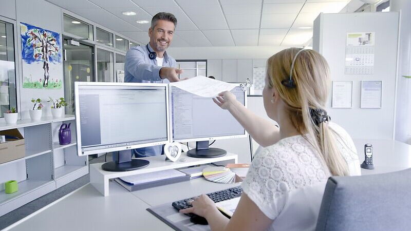 Ein Mann und eine Frau im Büro, die Frau sitzt am Schreibtisch, der Mann reicht ihr ein Dokument