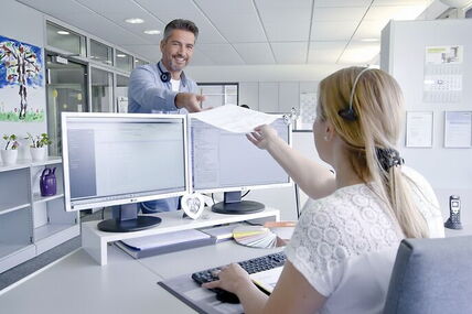 Ein Mann und eine Frau im Büro, die Frau sitzt am Schreibtisch, der Mann reicht ihr ein Dokument