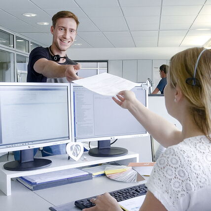 Ein Mann und eine Frau im Büro, die Frau sitzt am Schreibtisch, der Mann reicht ihr ein Dokument