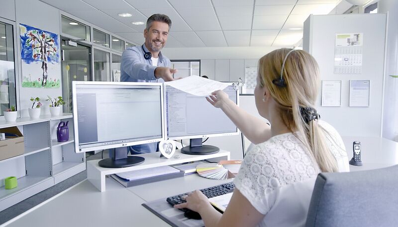 Ein Mann und eine Frau im Büro, die Frau sitzt am Schreibtisch, der Mann reicht ihr ein Dokument